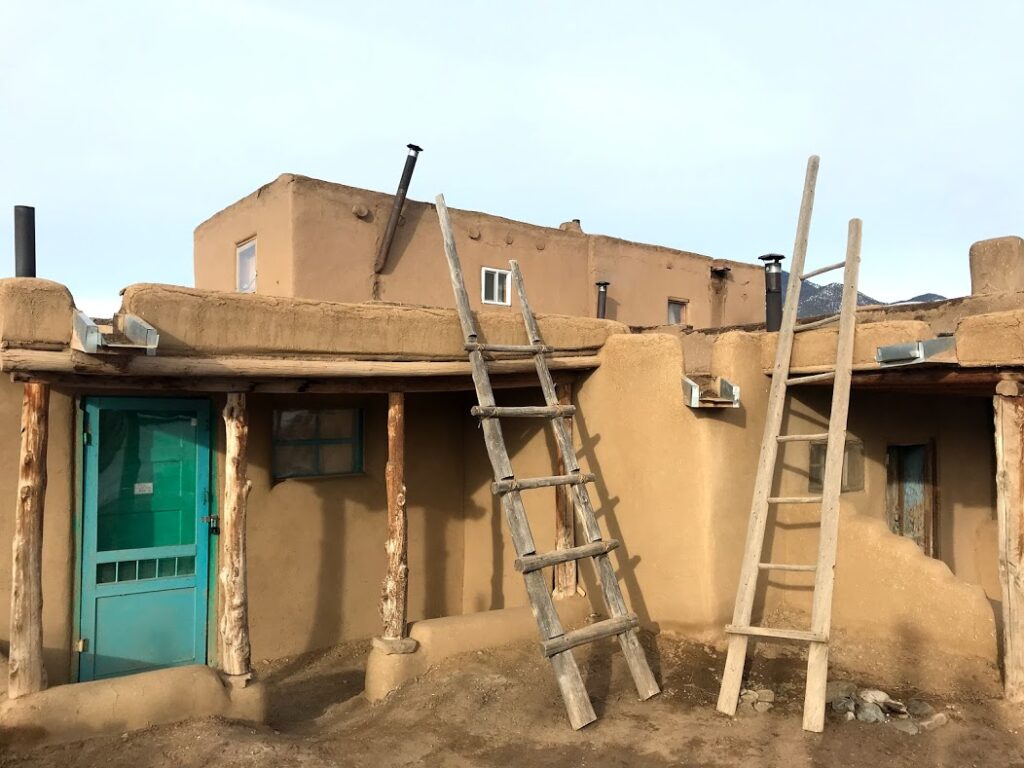 A two story adobe building with a turquoise door and wooden ladders leaning against it.