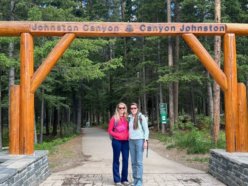 the author and friend stand under a large wooden sign that says Johnston Canyon on it as they prepare to go hiking in Johnston Canyon