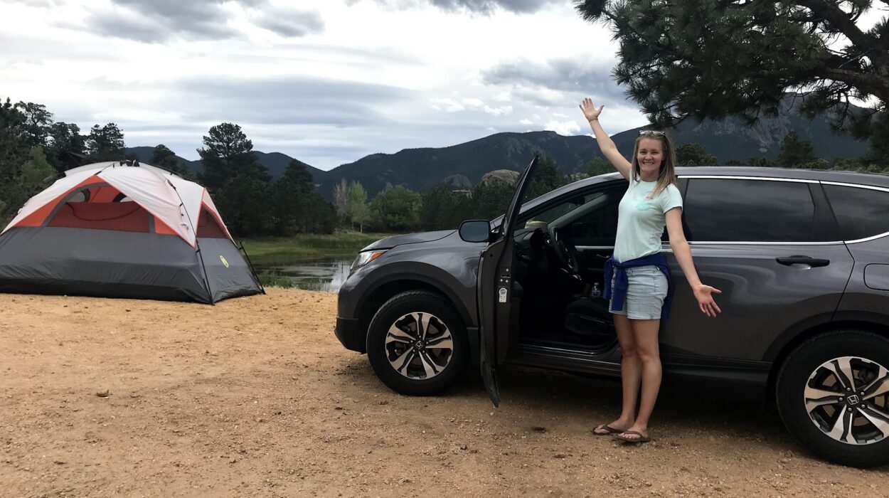 a girl stands outside of a car parked next to a tent at a campsite.