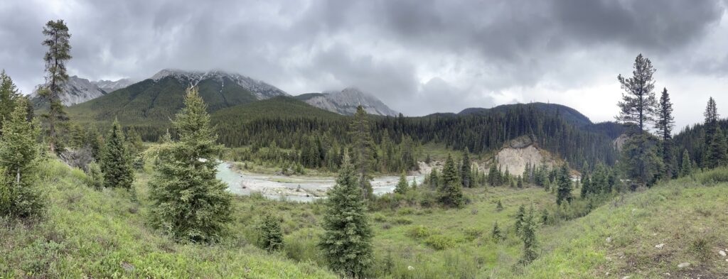 a panoramic view of gray skies and clouds over bare mountains, a flowing river, and various evergreen trees