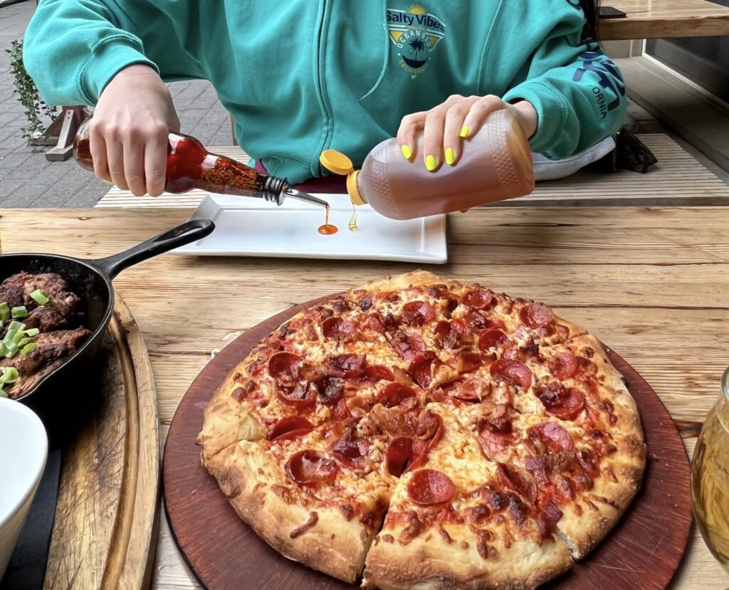 a delicious looking piece of pizza from Bear Street Tavern (one of the best places to eat in Canmore and Banff, Canada) sits in the foreground, while the author pours honey and homemade rosemary chili oil onto a plate in the background