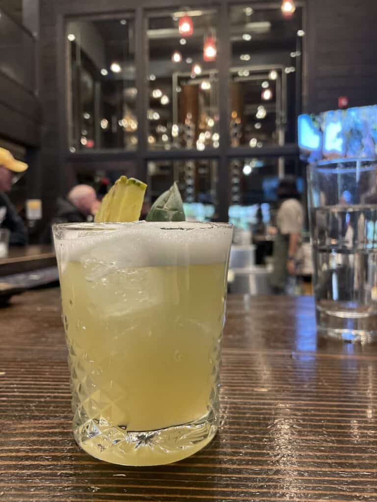 a small cocktail witha pineapple garnish sits on a table with the Park Distillery restaurant in Banff appears in the background 