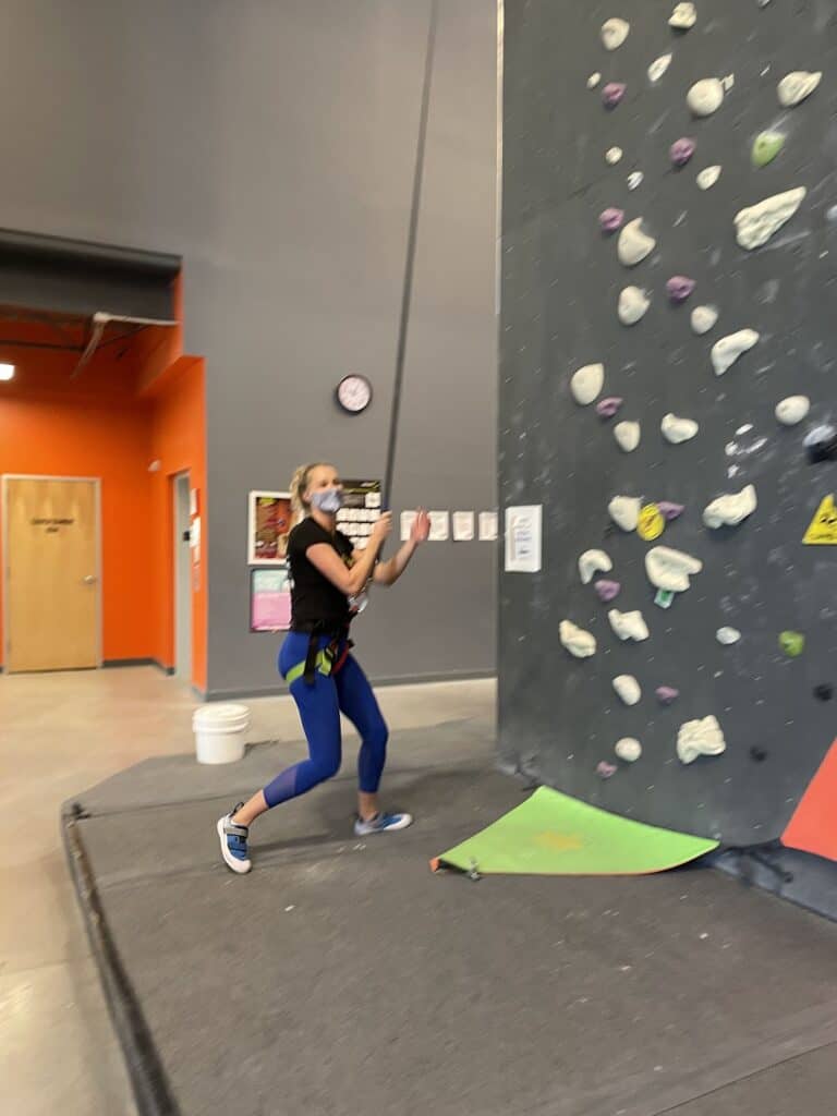 a blond woman landing after rappelling down a wall in an indoor rock climbing gym 