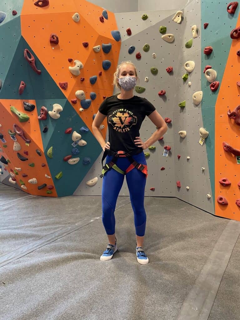 the author proudly stands in her climbing harness and velcro rock climbing shoes in front of the indoor rock climbing wall 