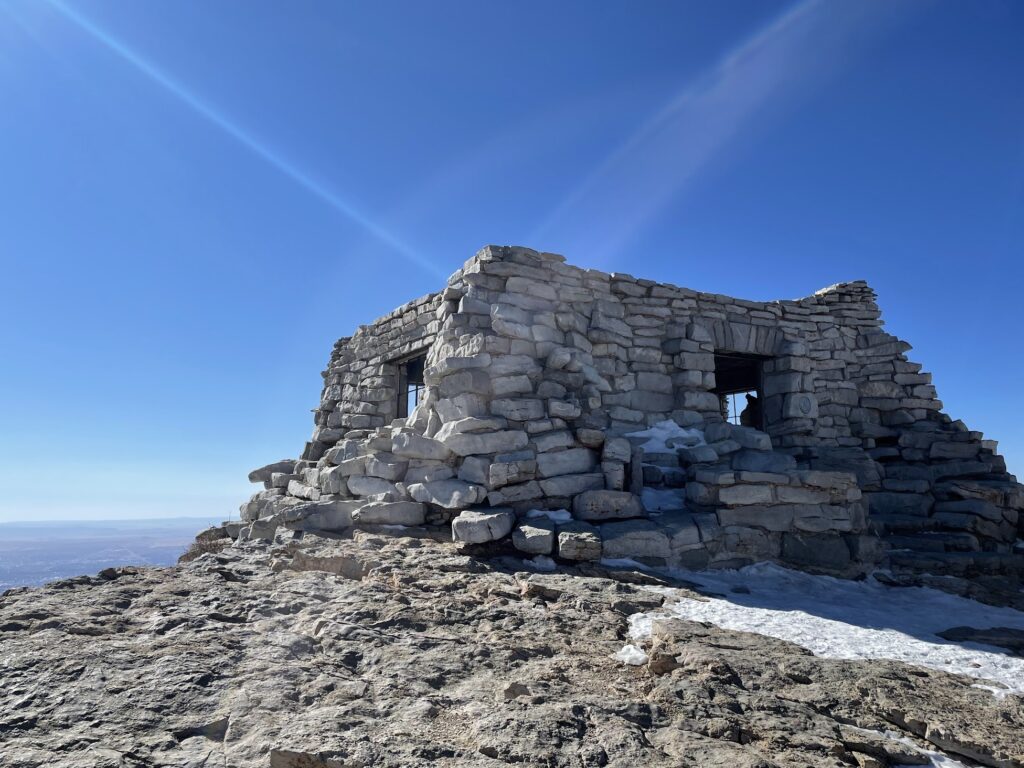 The kiwanis cabin, a small rock cabin perches on the edge of the Sandia mountain with blue sky in the background.