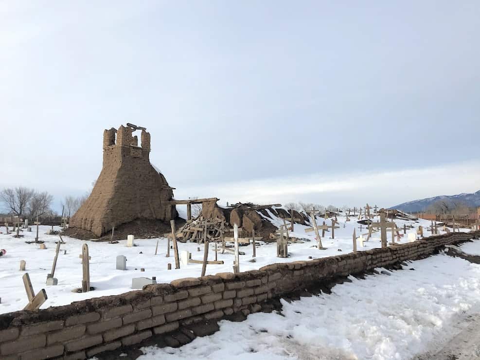 The remains of an adobe church. There is a small adobe brick wall on the border, snow covers the ground of the graveyard, and only the adobe belltower remains.