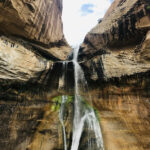 a waterfall pours down a tan rockface, splitting into a separate stream of water at Lower Calf Creek Falls in Utah