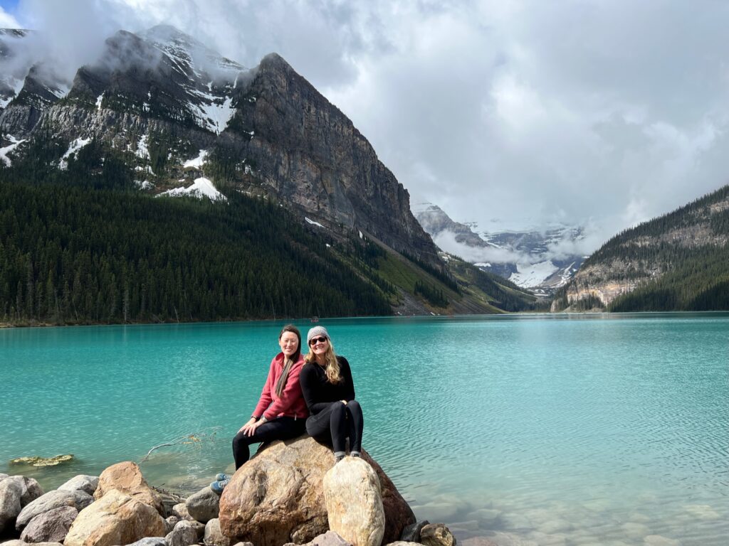 two women sitting on a rock smiling at the camera with Lake Louise and mountains in the background 