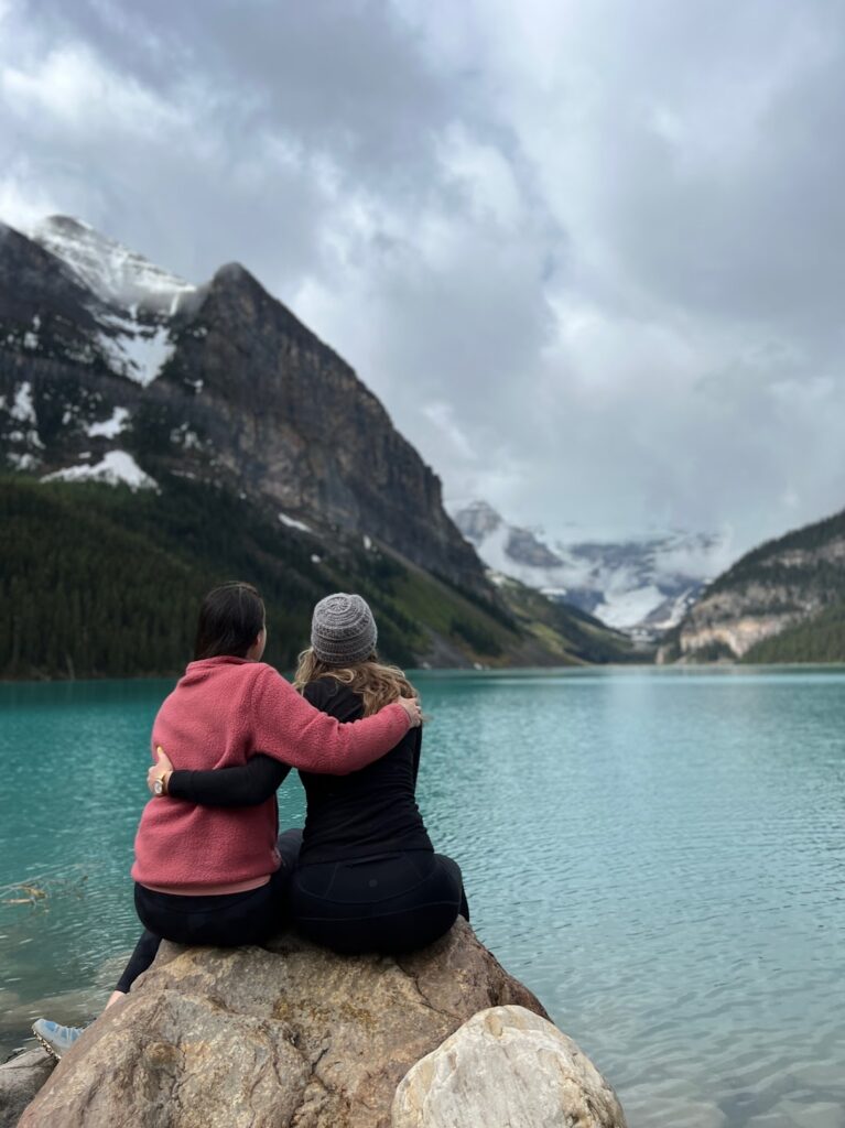 two women sit on a rock wit their arms around each other. Their backs are to the camera, and they are facing the turquoise water of Lake Louise 