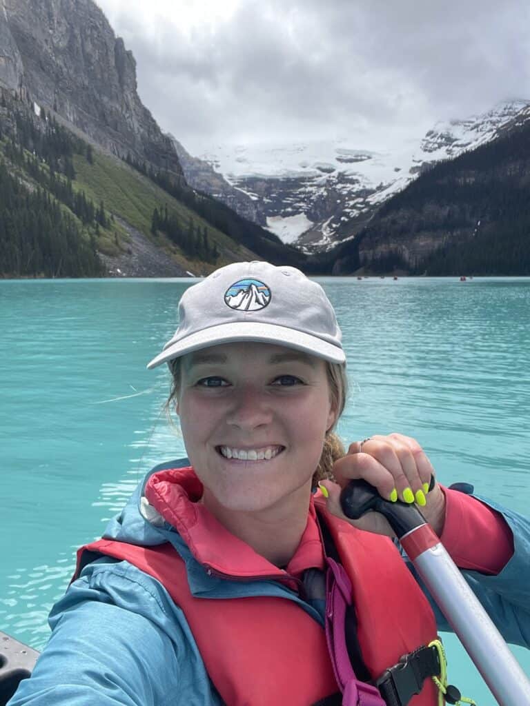 the author is holding a canoe paddle while smiling at the camera from inside a canoe on Lake Louise. Snow and mountains are also in the background.