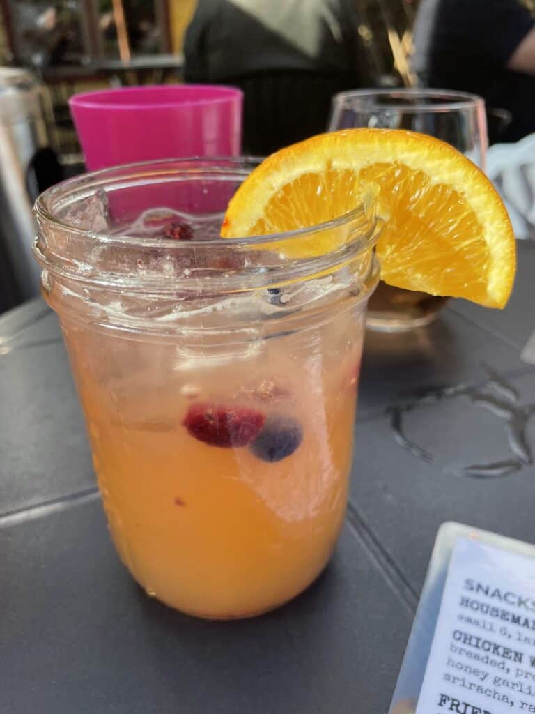 A mason jar with white peach sangria from tavern 1883, garnished with an orange slice