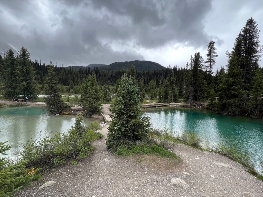 two bright turquoise pools and one light green pool glimmer under cloudy skies. A path with a small bridge cuts over the pools.