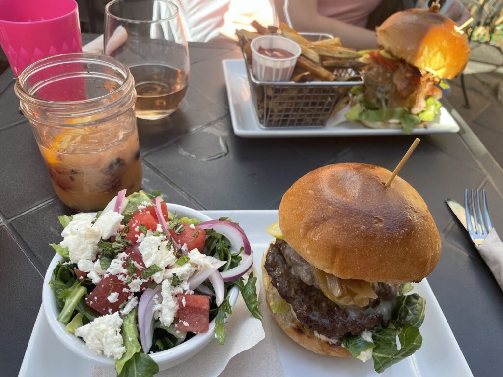 two decadent burgers from Tavern 1883, one of the best places to eat in Canmore and Banff, Canada. The burgers are perched atop rectangular white plates. There is also a watermelon, feta and mint salad, as well as a half-drank glass of white peach sangria