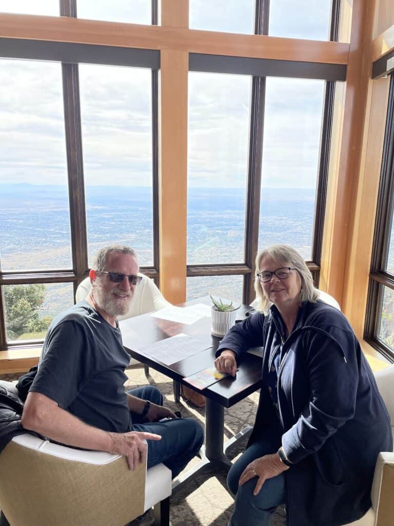 the author's mom and dad smile while seated at a table at the Ten 3 restaurant, with an incredible view of Albuquerque behind them out the beautiful floor to ceiling windows in the restaurant