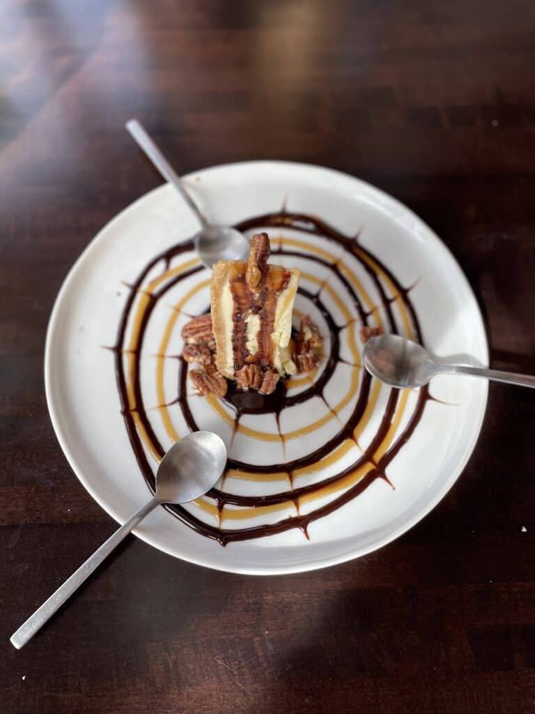 a delicious piece of cheesecake topped with pecans sits atop a plate drizzled with a caramel and chocolate sauce spiderweb design. Three spoons rest on the plate