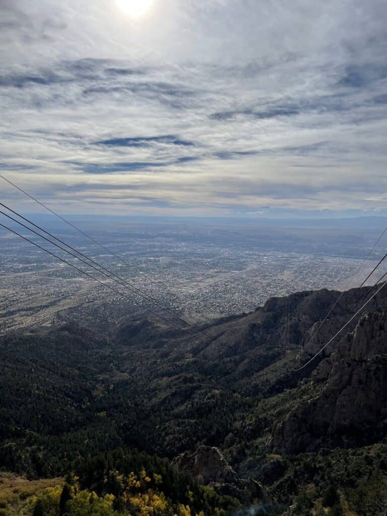the wires from the Sandia Peak tram descend down the mountainside, with a hazy sky in the background and an incredible aerial view of Albuquerque from the tram