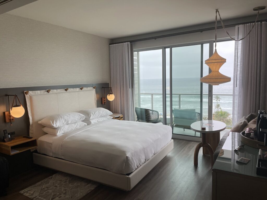 a white bed in a well-designed room at the Mission Pacific Inn sits in front of glass doors that show a floor to ceiling ocean view 