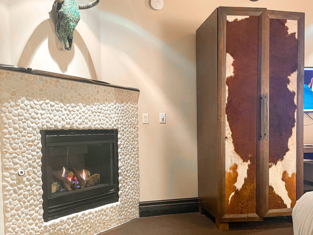 a white stone fireplace, lit with a fire, underneath a turquoise-decorated skull, next to a cowhide armoir in the Taos Mountain room at El Monte Sagrado 