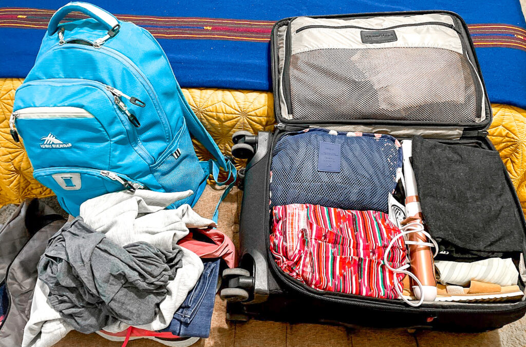 a suitcase lies open, displaying packing cubes inside and other clothes. a backpack is next to the suitcase, as is a pile of clothing