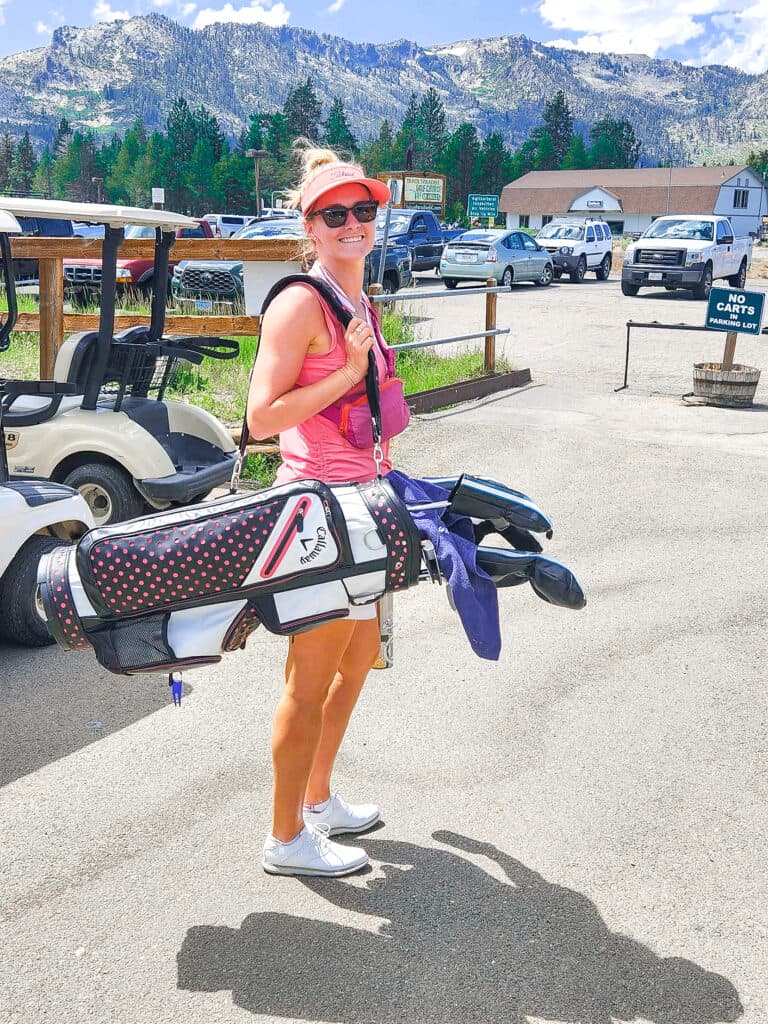 the author stands in the foreground holding her golf clubs with mountains in the background 