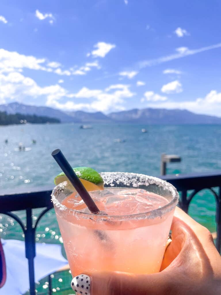 a pink paloma drink  from the Boathouse Pier is in the foreground with Lake Tahoe's sparkling blue waters and mountains in the background